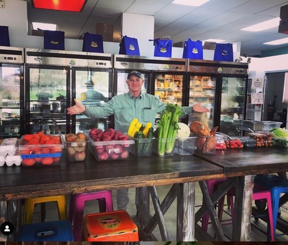 Man with arms raised in kitchen with fresh produce in bins.