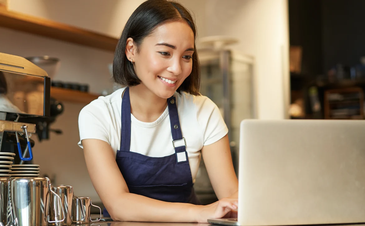 Restaurant operator wearing an apron uses a laptop at a counter beside an espresso machine.