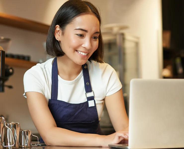 Restaurant operator wearing an apron uses a laptop at a counter beside an espresso machine.