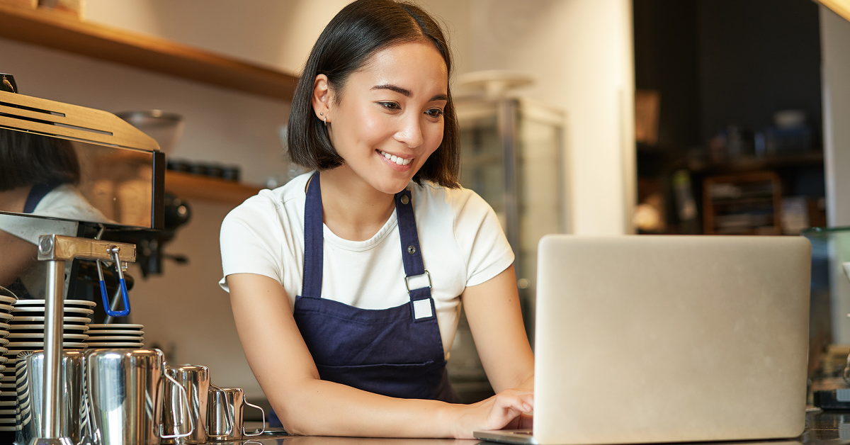 Restaurant operator wearing an apron uses a laptop at a counter beside an espresso machine.