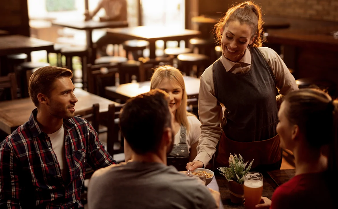 Restaurant server delivers food to four guests seated around a table in a warmly lit dining room.