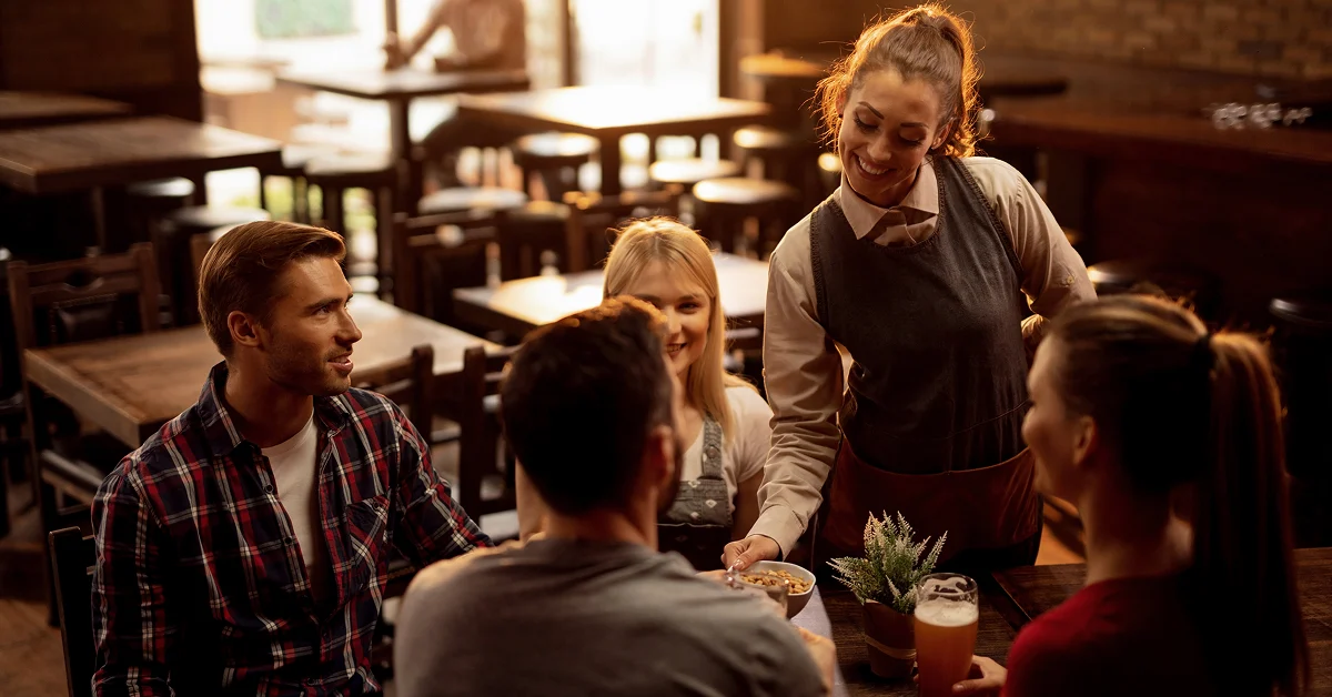 Restaurant server delivers food to four guests seated around a table in a warmly lit dining room.